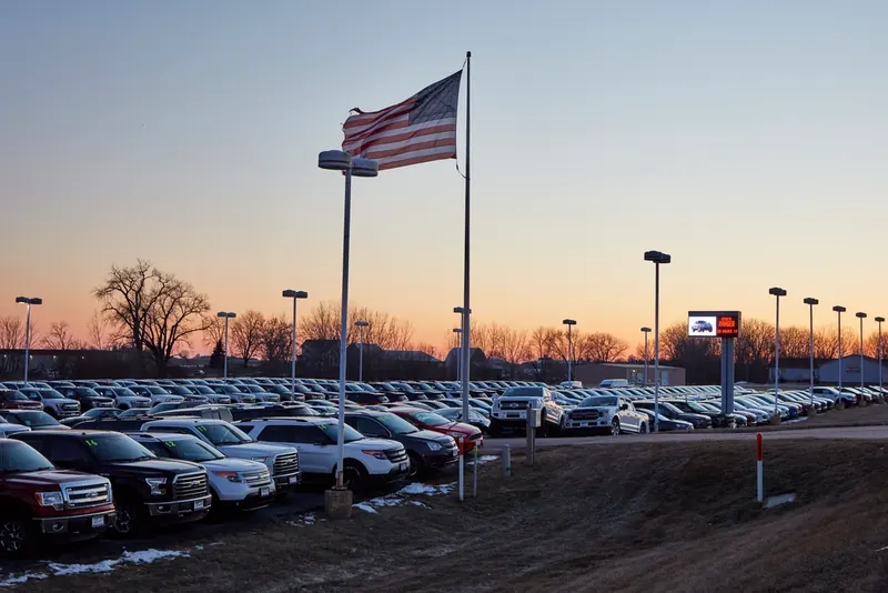 Large car dealership lot full of trucks and SUVs at dusk under an American flag