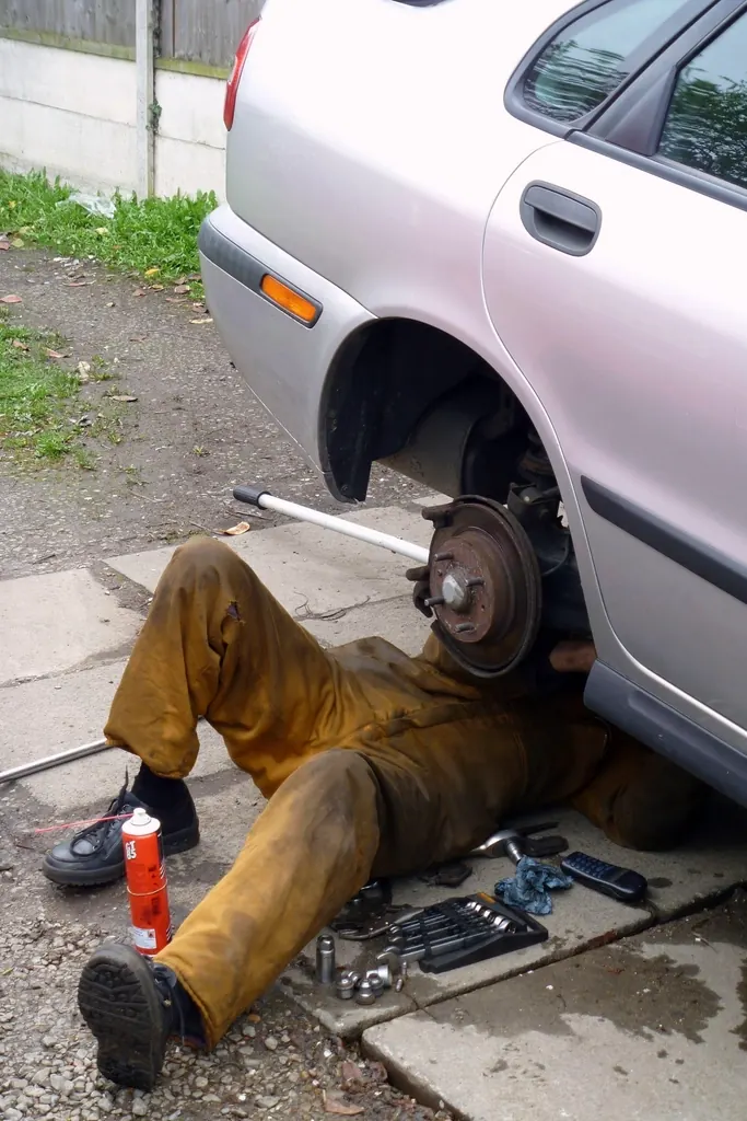 Mechanic in work pants lying under a silver car with a wheel removed and tools nearby