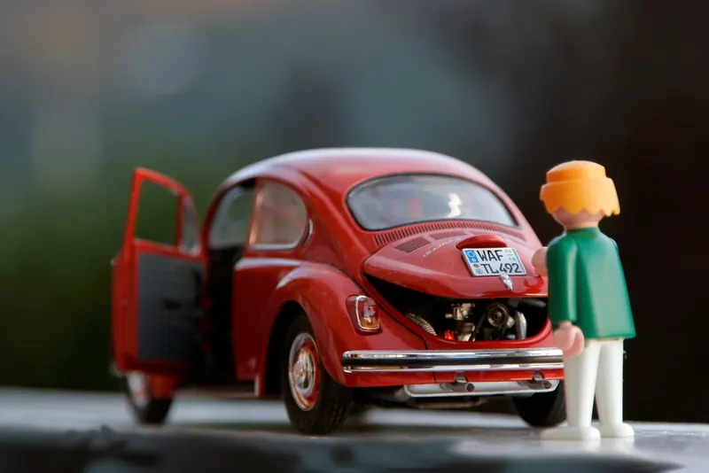 Toy figure standing beside a red toy Beetle with its doors and engine lid open