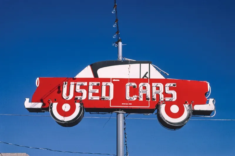 Red car-shaped USED CARS neon sign on a pole against a deep blue sky